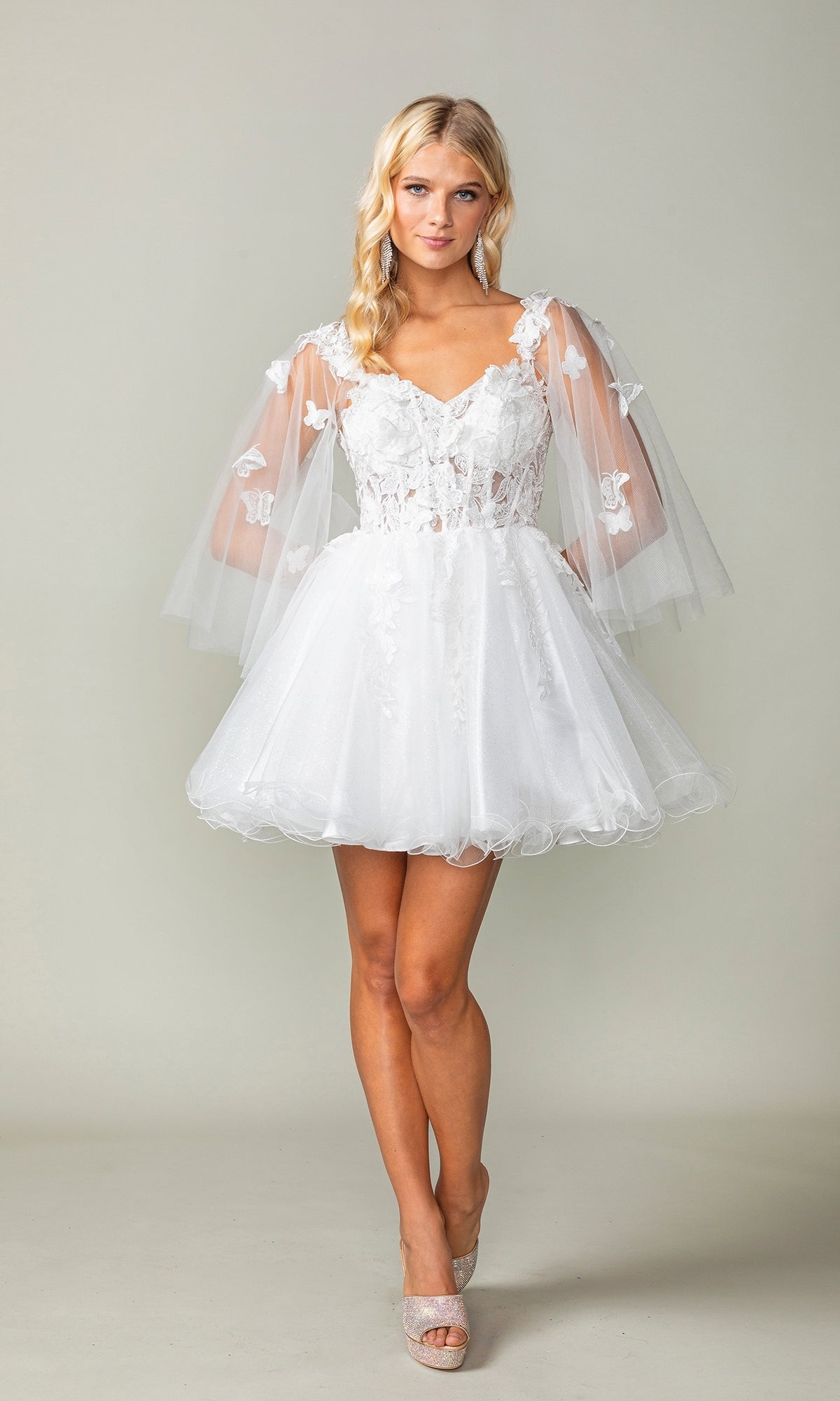 A woman wears the Butterfly-Cape Short A-Line Homecoming Dress 3393 with sheer, flared sleeves, lace floral details, and a tulle skirt, standing against a plain light background.